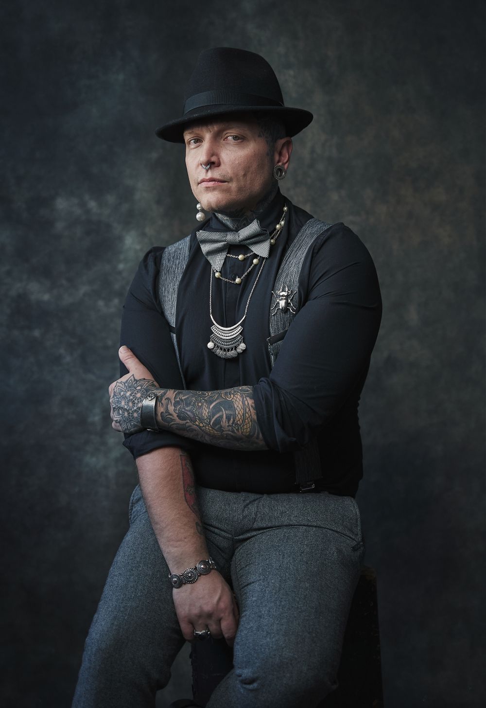 Tattooed man wearing a black hat, suspenders, and bow tie poses against a dark textured background.