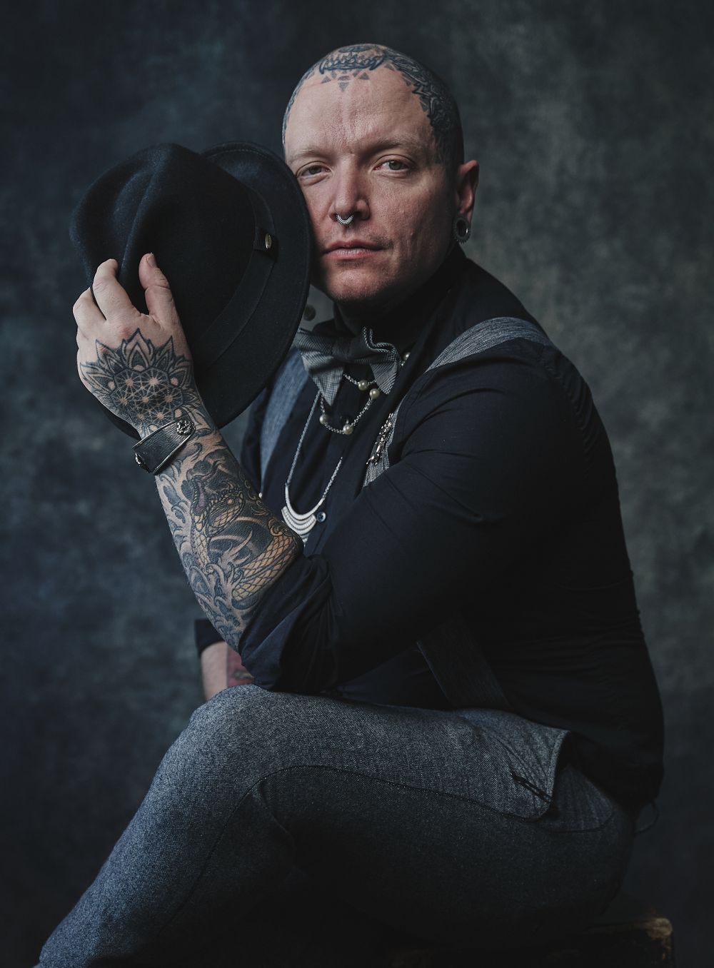 Tattooed man with shaved head holding black hat in dramatic studio portrait.