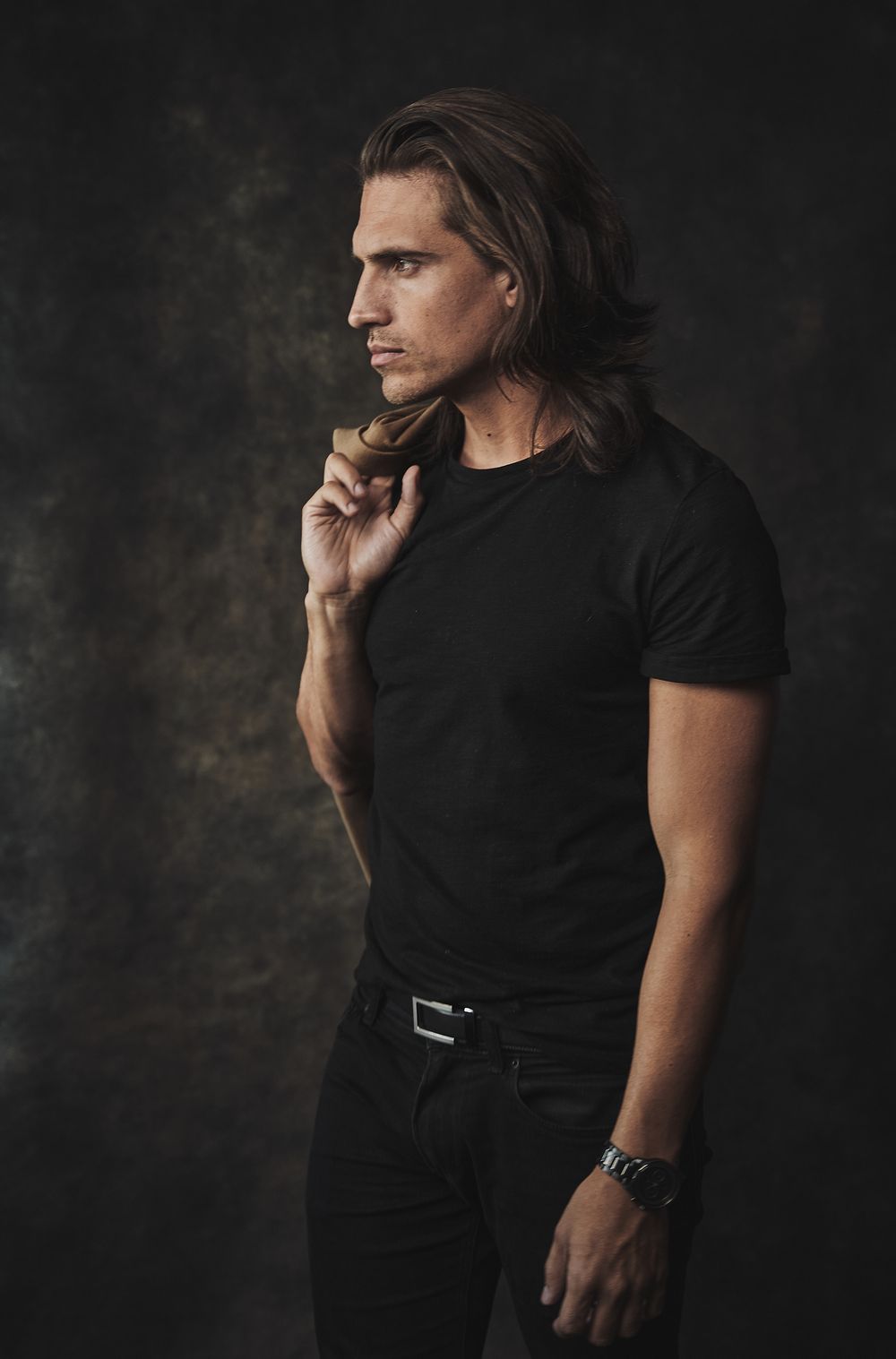 Athletic man with long hair in black outfit holding pipe against dark textured background.