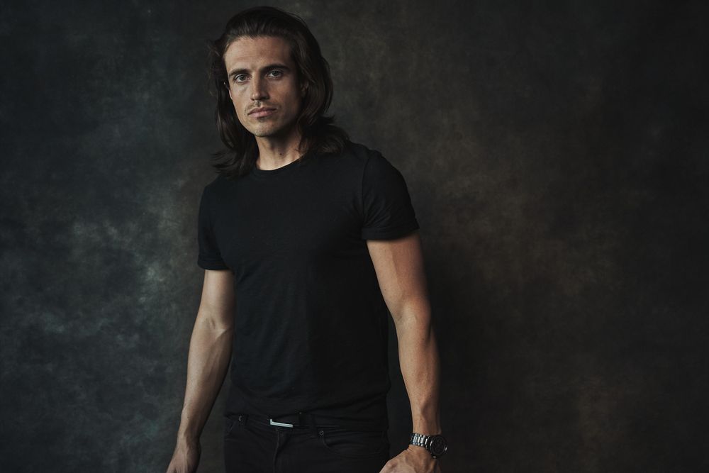 Man in black t-shirt with long dark hair poses confidently against a dark textured studio background.