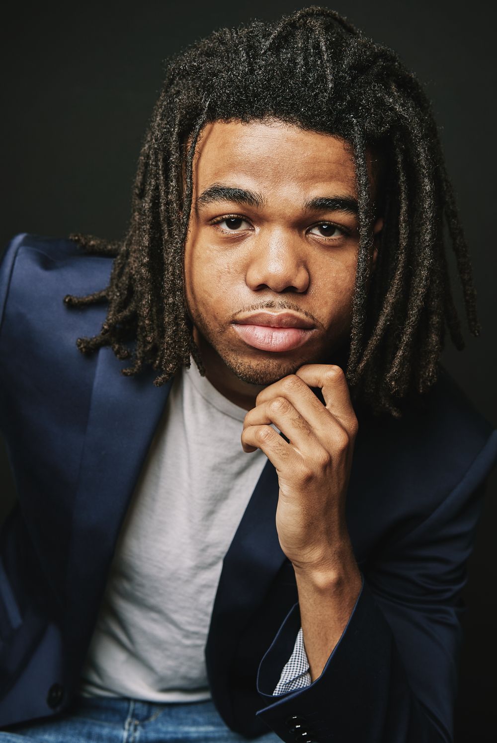 Young man with dreadlocks in navy blazer with chin resting on hand.