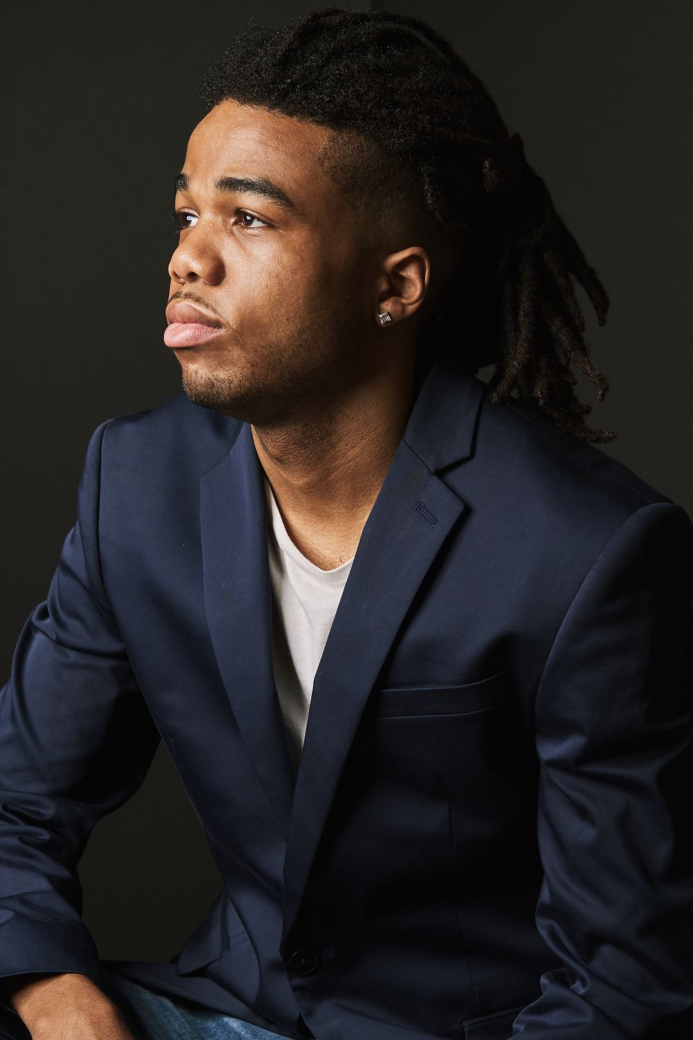 Young Black man with dreadlocks wearing a navy blazer sits against a dark background looking away.