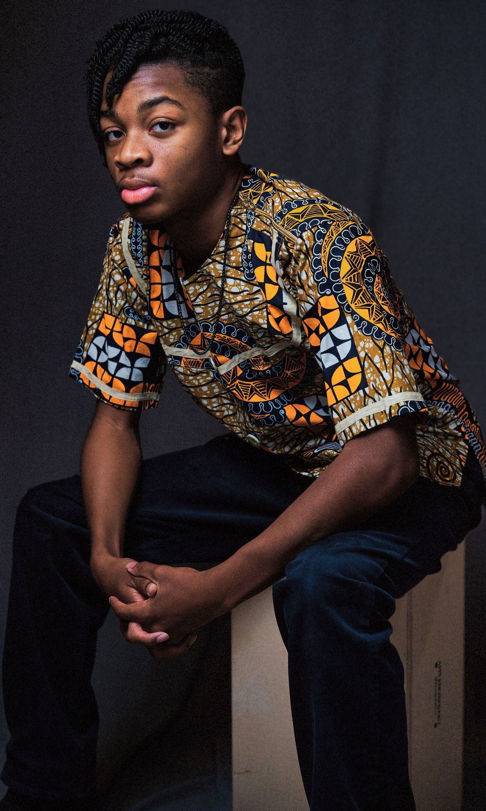 Young man in colorful African print shirt sitting on box against dark background.