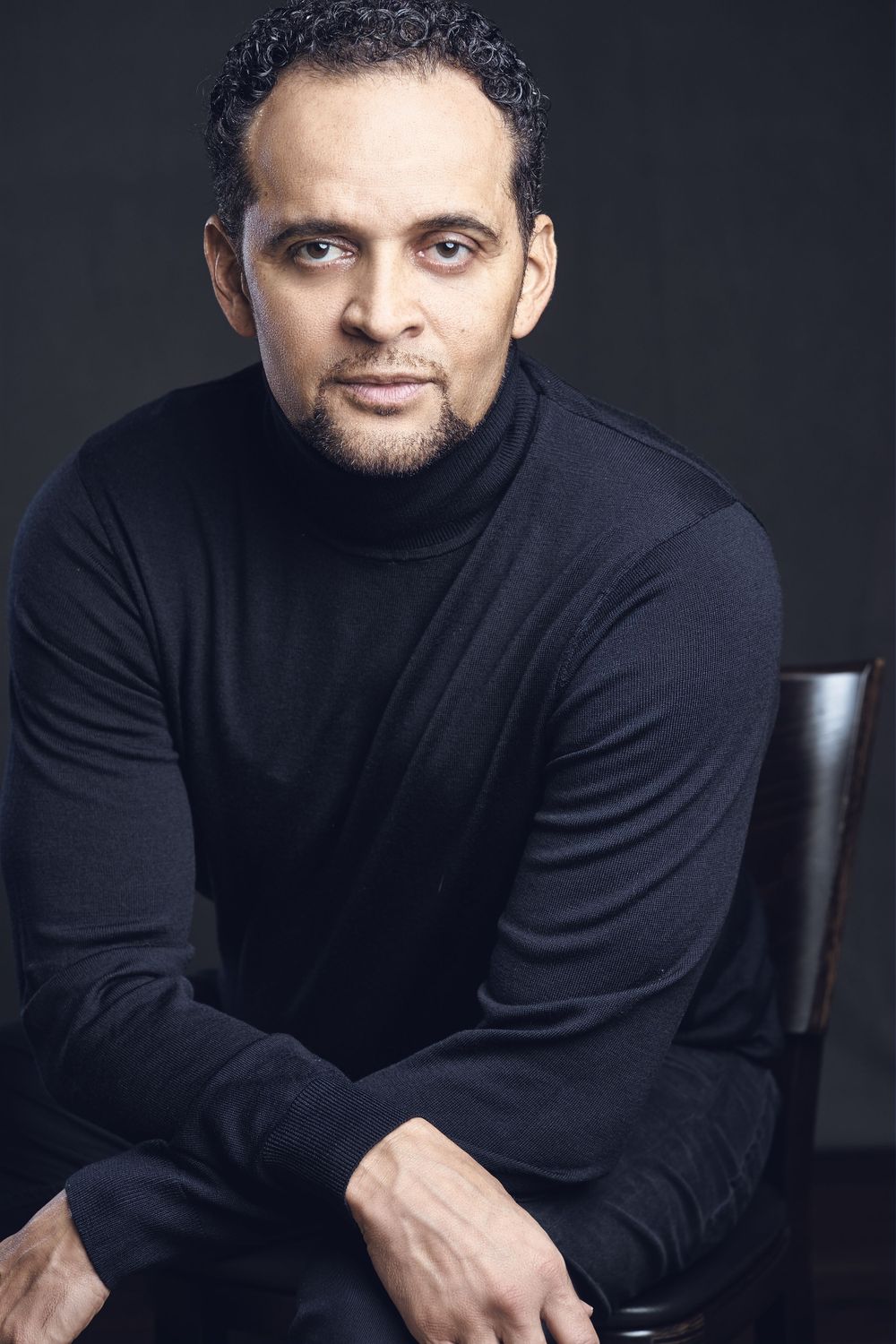 Man in black turtleneck seated in chair against dark background in studio portrait.