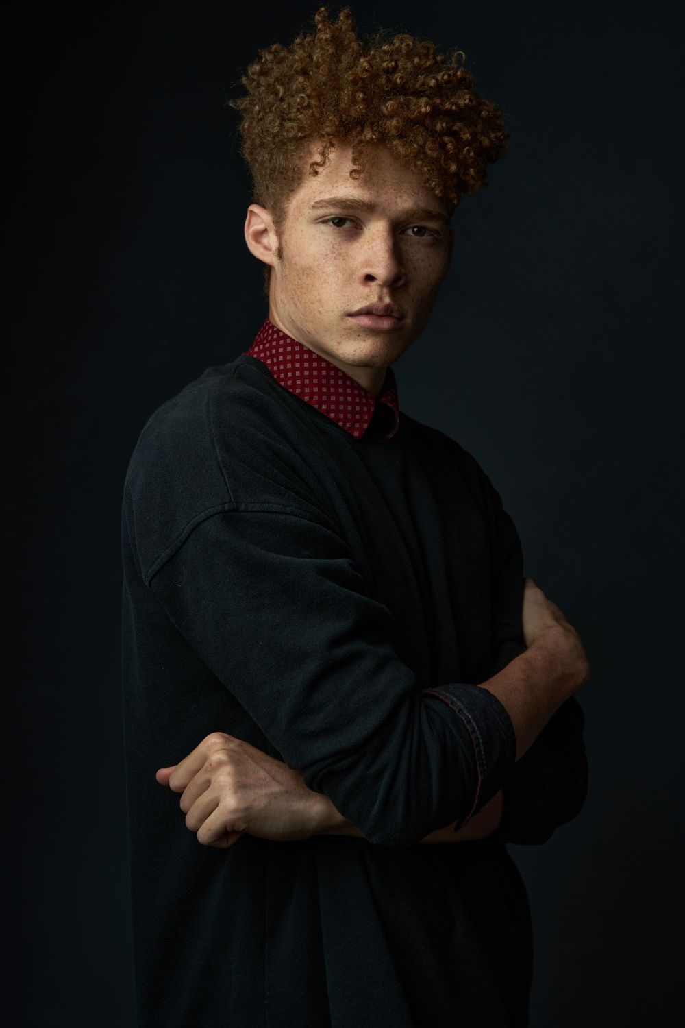 Young man with curly auburn hair and freckles poses with arms crossed in a dark studio setting.
