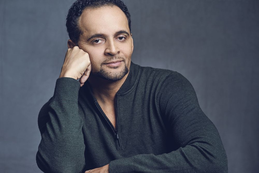 Man in gray sweater resting chin on hand with soft smile against gray background.