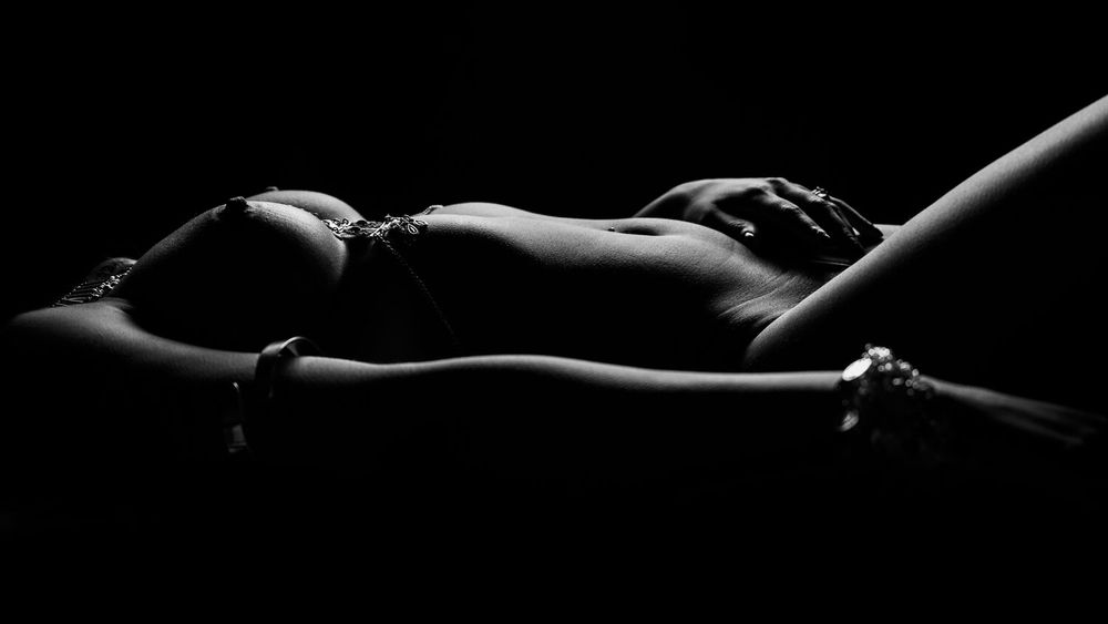 Black and white artistic nude photograph showing dramatic shadows and curves of a reclining silhouette against dark background.