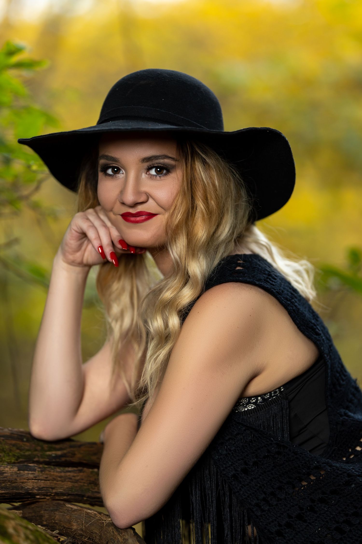 Person in black outfit and wide-brimmed hat poses outdoors with autumn backdrop.