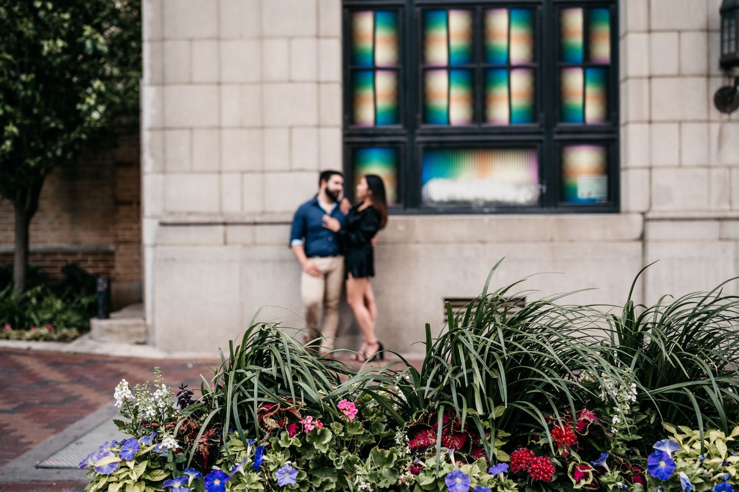 A couple stands together by a decorative window while colorful flowers bloom in the foreground.