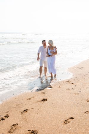 A couple dressed in white walks hand in hand along the beach shoreline leaving footprints in the sand.
