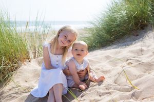 Two people sit together on sandy dunes with beach grass and ocean waves in the background.