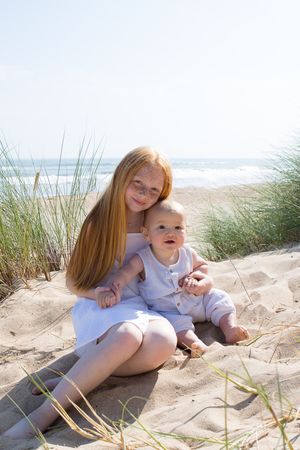 A parent and baby sit together among beach grass dunes on a sunny day.