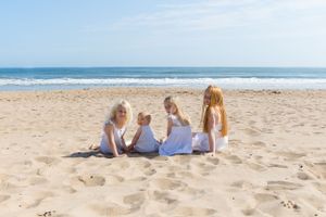 Family members wearing white dresses sit together on a sunny beach near the ocean.