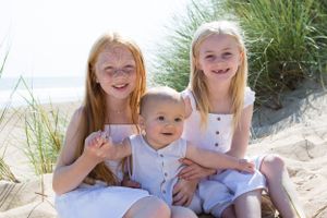 Three people in white dresses sit together smiling on a sandy beach with dune grass.