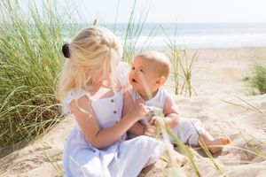 Two figures in white dresses share a tender moment among beach grass on a sunny seashore.