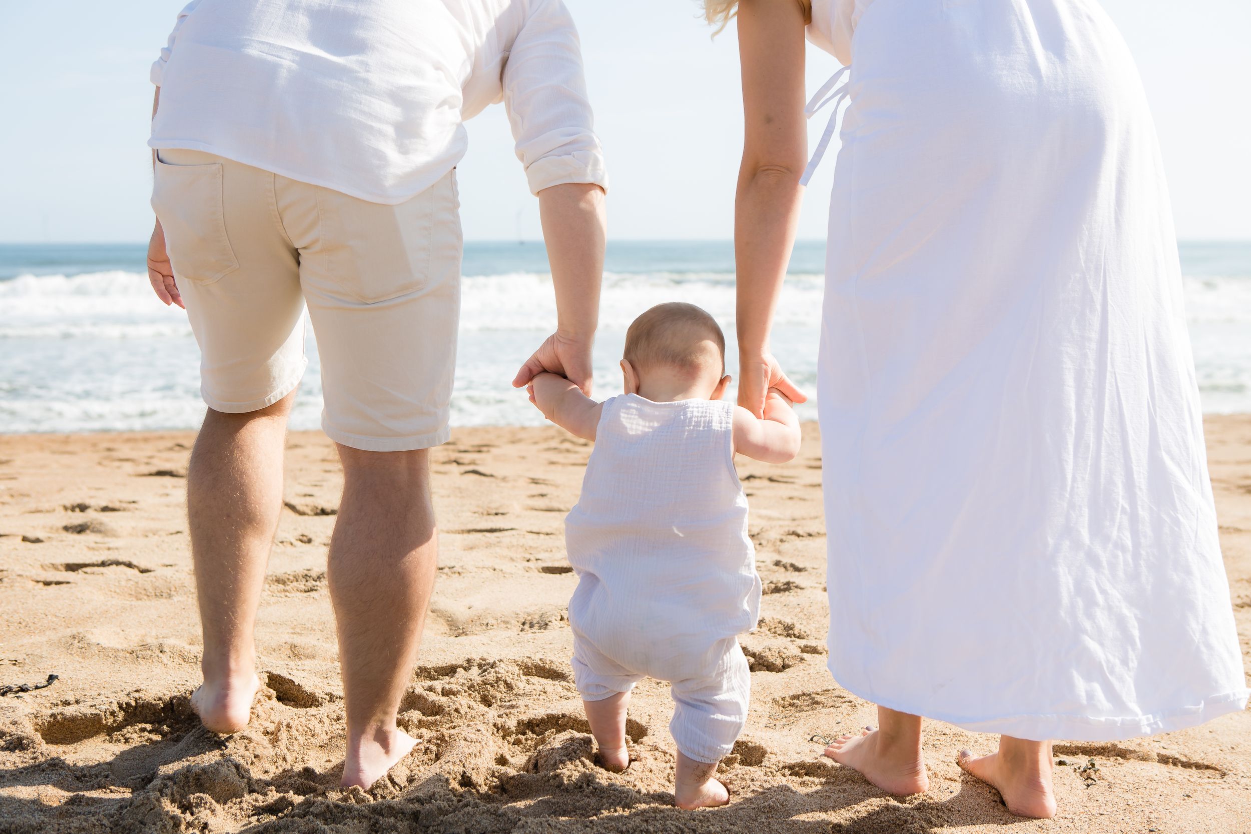 Family walking together holding hands on sunny beach with toddler in white outfit.