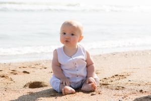 A baby in white clothing sits on sandy beach with ocean waves in background.