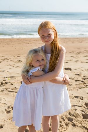 Two people in white dresses embrace on a sunny beach with waves in the background.