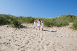 People in white clothing walk up sandy dunes with green grass on a bright summer day.