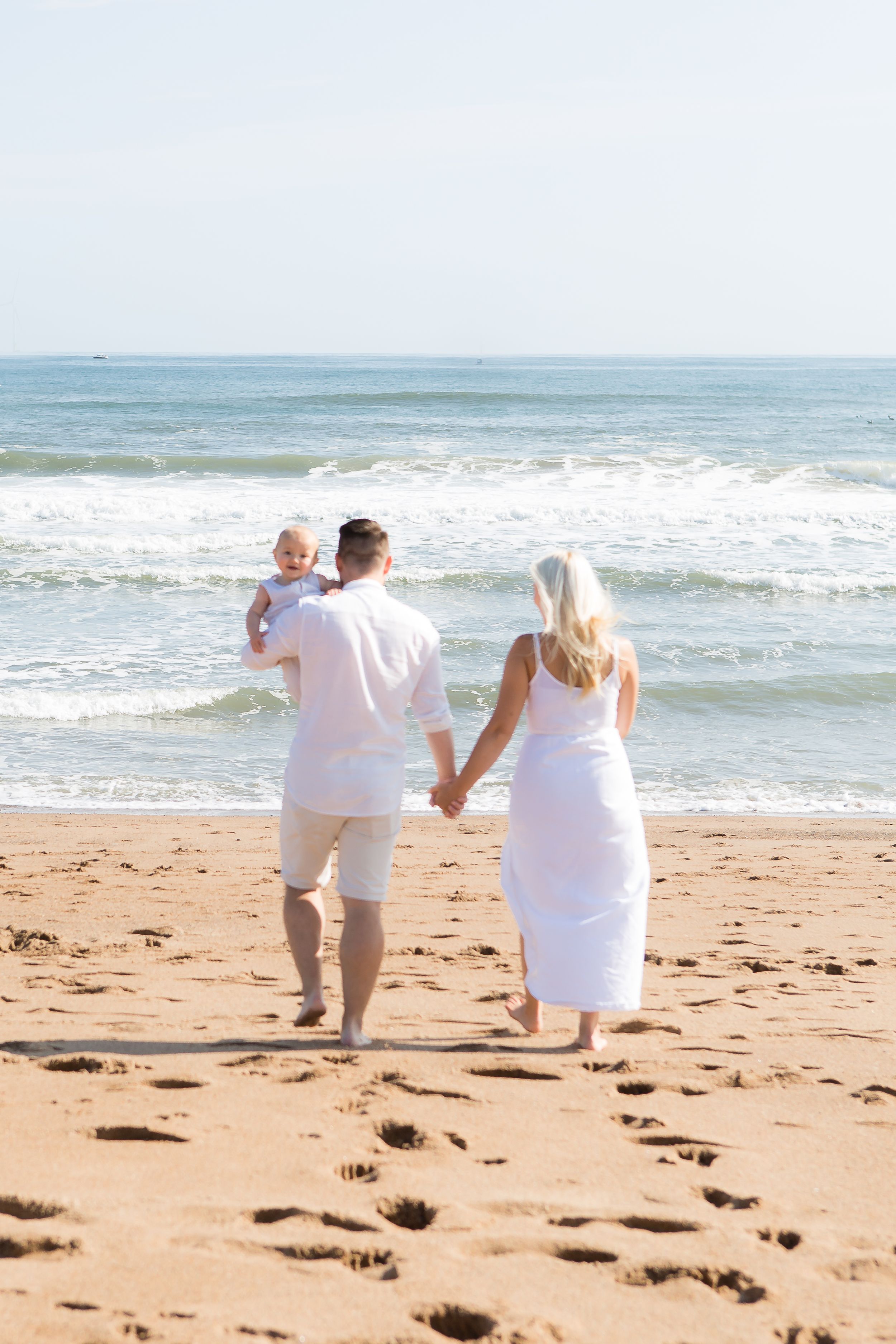 Family in white clothing walking along beach shoreline leaving footprints in sand.