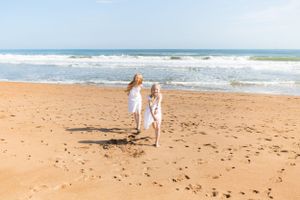 Two people in white dresses walk hand-in-hand along a sandy beach.