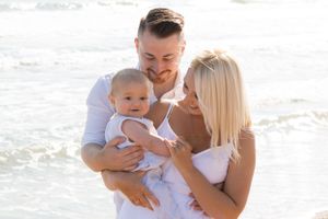 A happy family dressed in white embraces while standing on a beach with ocean waves in the background.