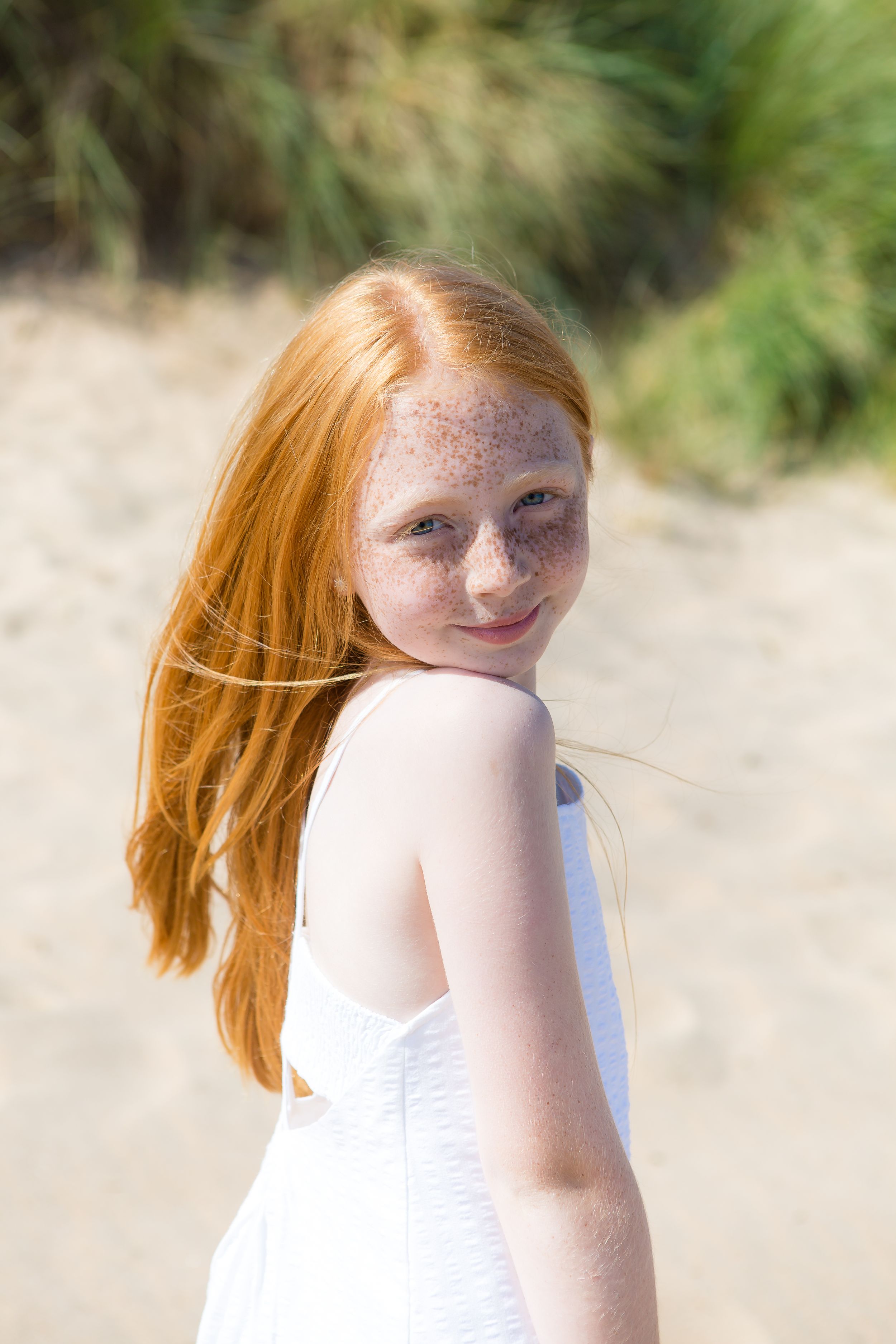 Person in white dress with flowing hair poses on beach with dunes in background.