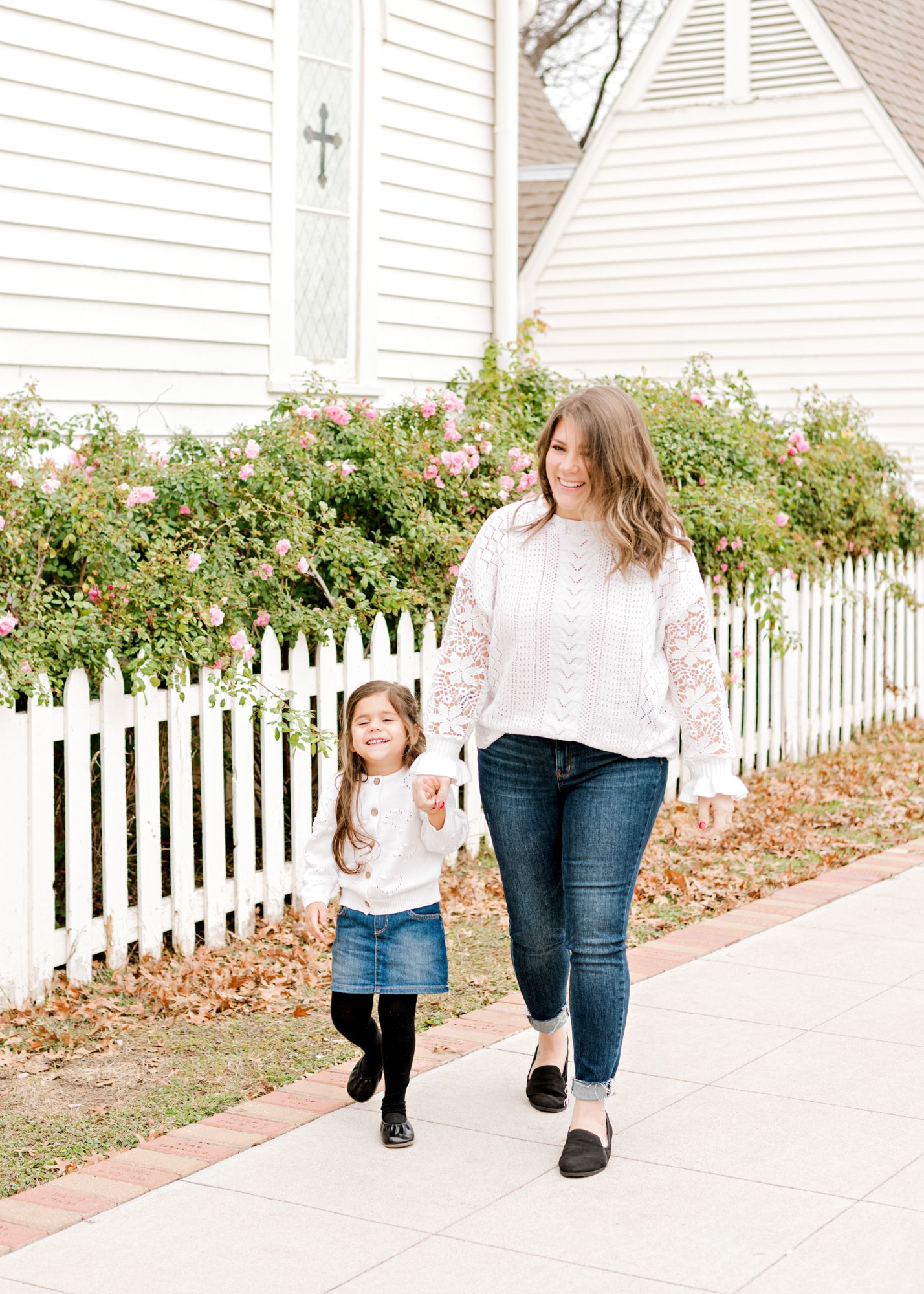 Mother and daughter wearing matching white sweaters walk together along a sidewalk beside a white picket fence and pink roses.
