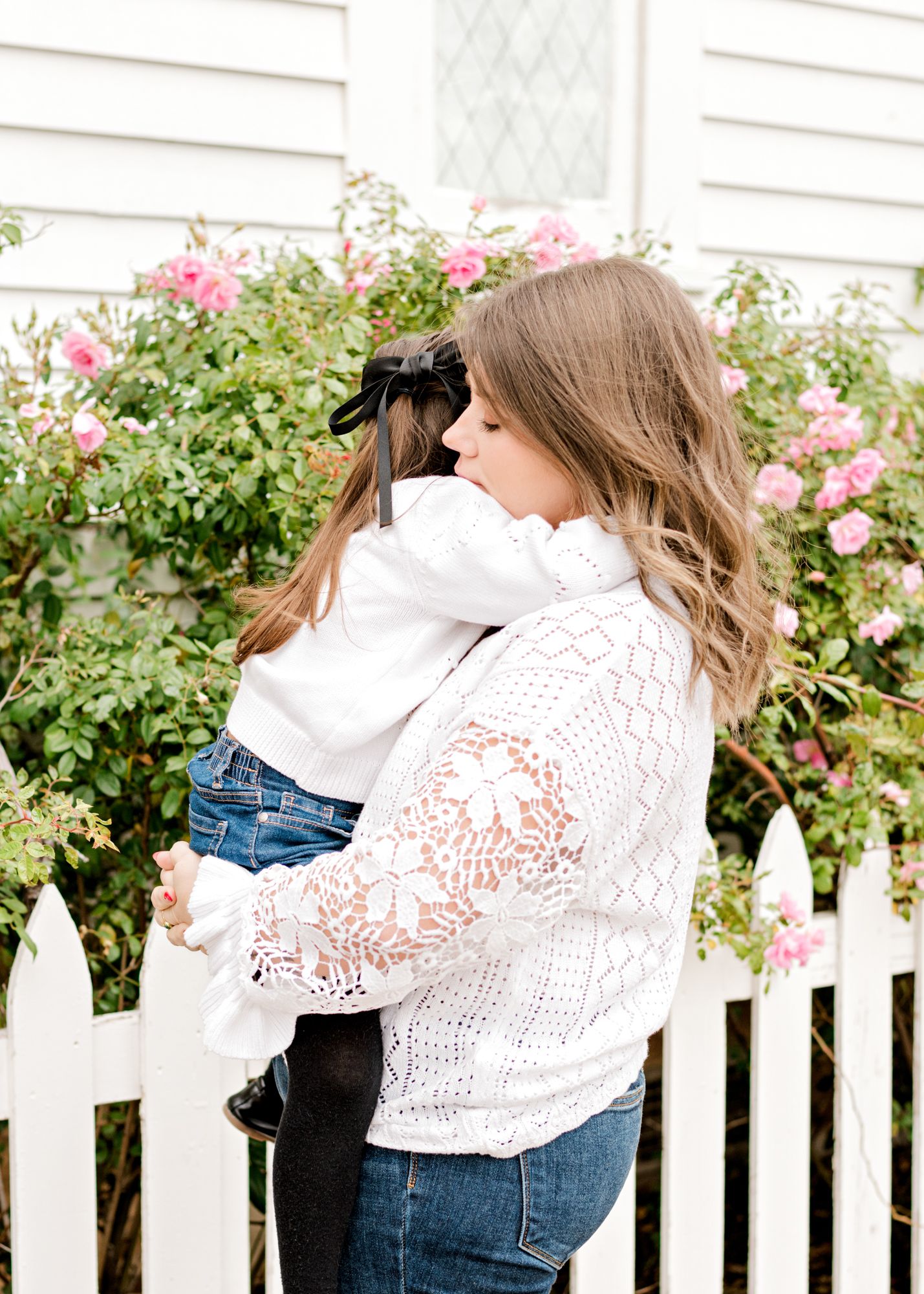 Mother and daughter in a sweet embrace by pink rose bushes and a white picket fence.