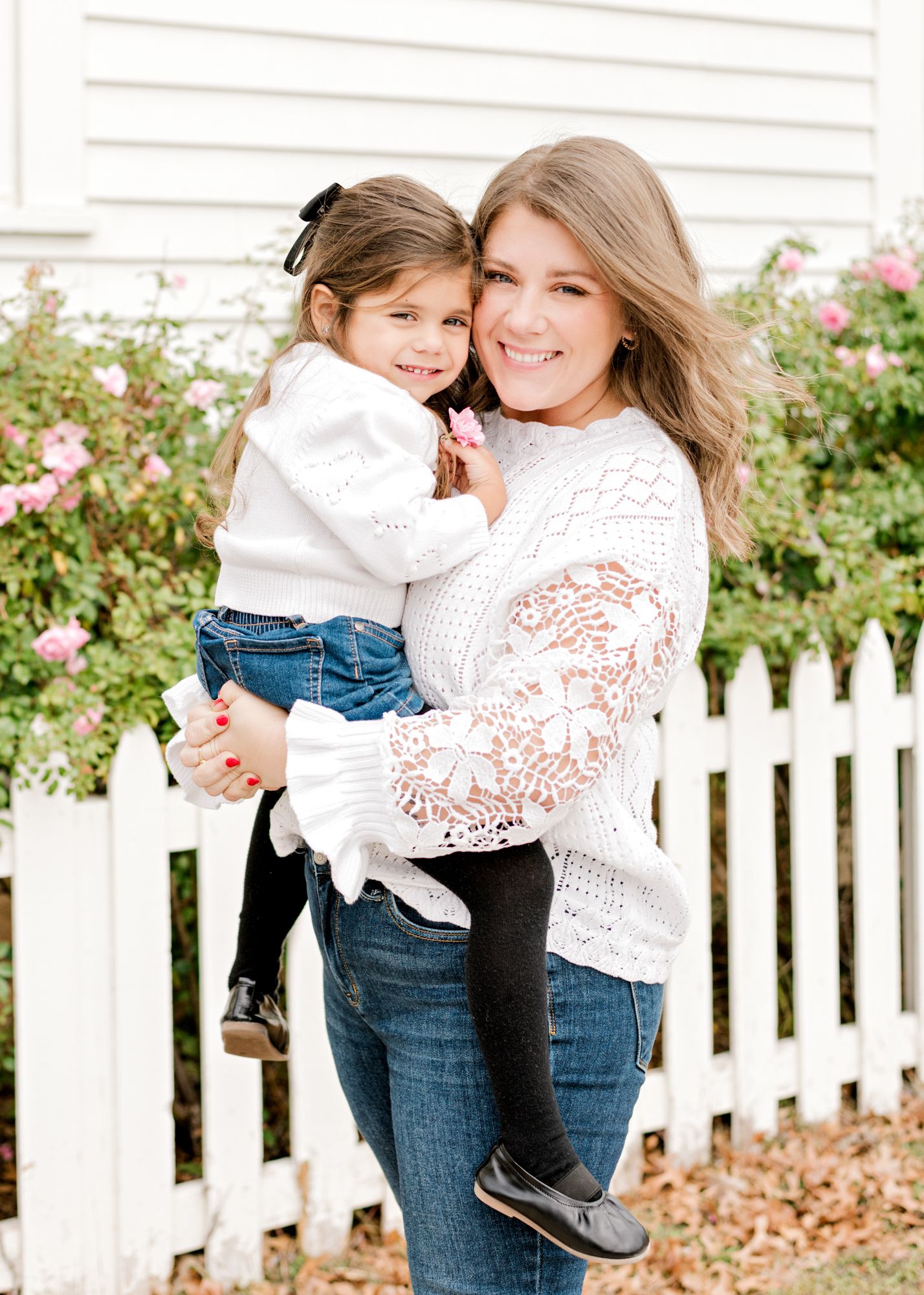 Two people in matching white sweaters share joyful moments by pink roses and a picket fence.