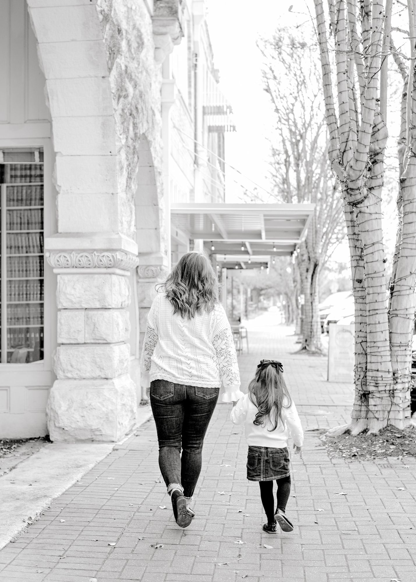 Two people hold hands while walking along a tree-lined brick pathway in black and white.