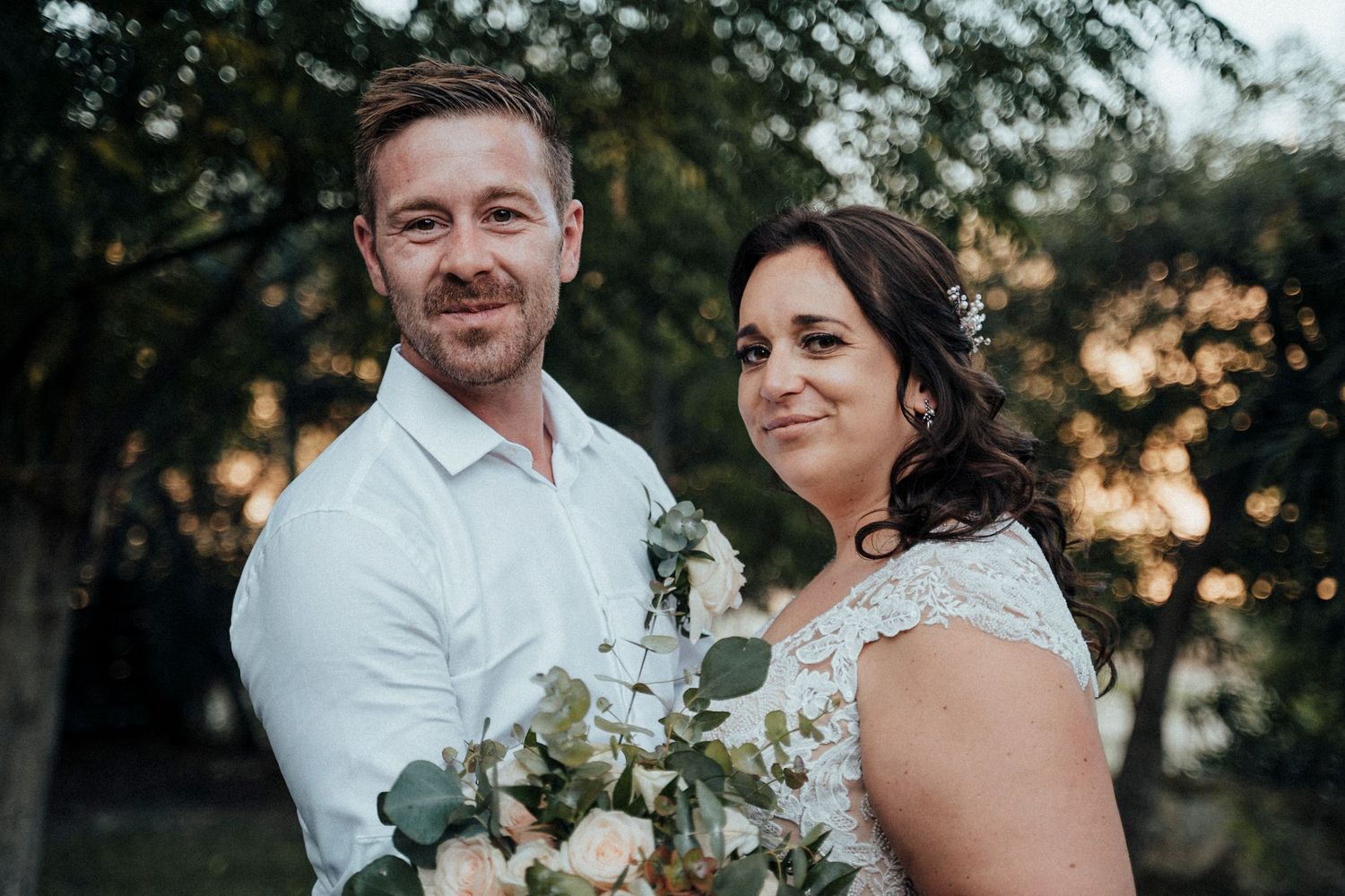 A couple poses with a white floral bouquet during their outdoor wedding photoshoot.