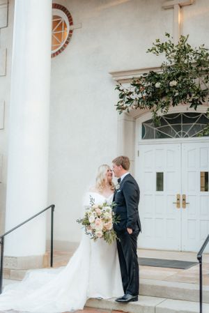 Bride and groom standing together outside classic white church, bride holding soft pink floral bouquet. Megan Snell Photo Utah Weddings