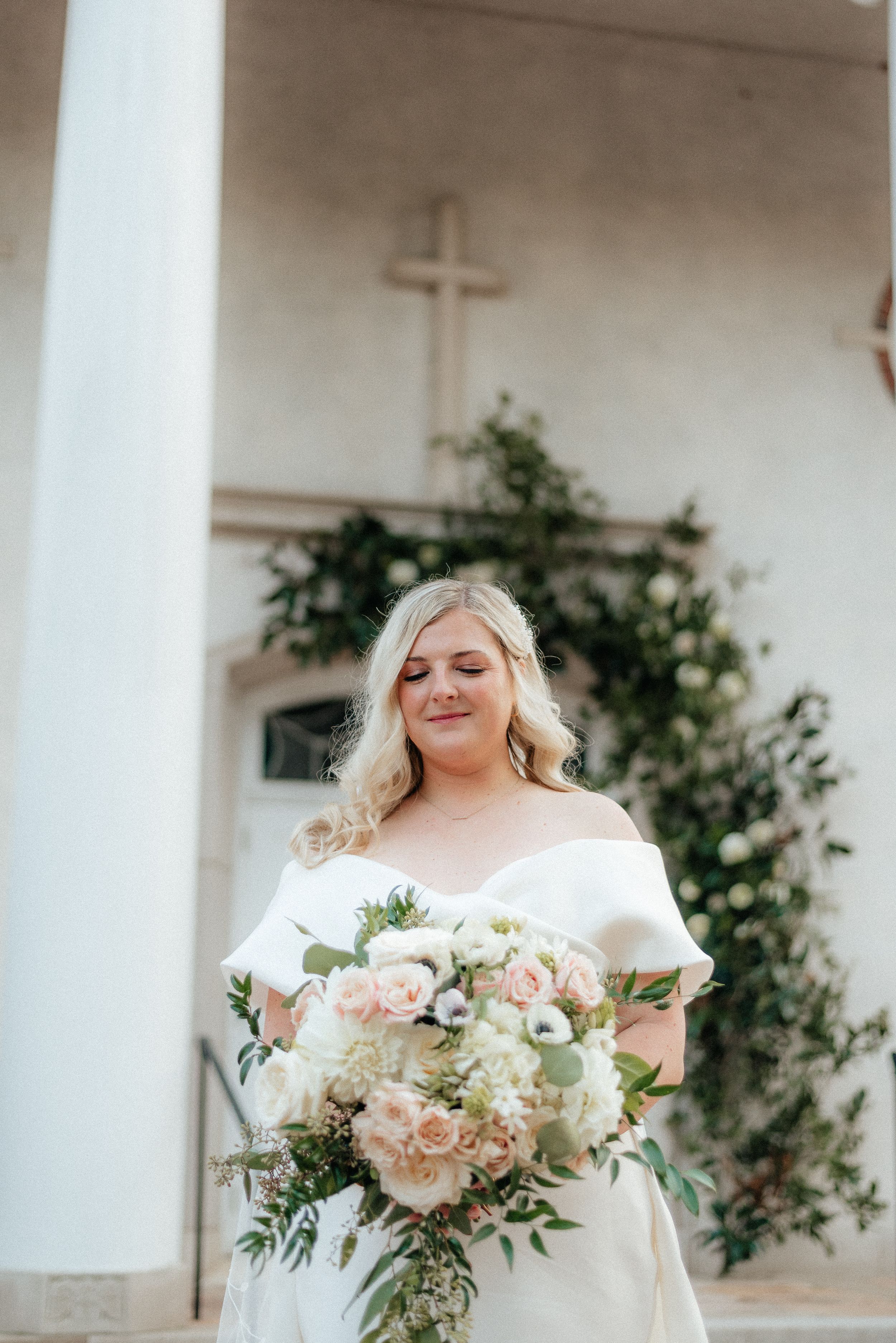 Bride in off-shoulder white gown holding blush and white bouquet stands before a cross-adorned church wall. Megan Snell Photo Utah Weddings