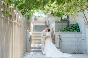 Bride and groom share a kiss in a sunlit outdoor corridor with lush trees and stone steps at their wedding venue. Megan Snell Photo Utah