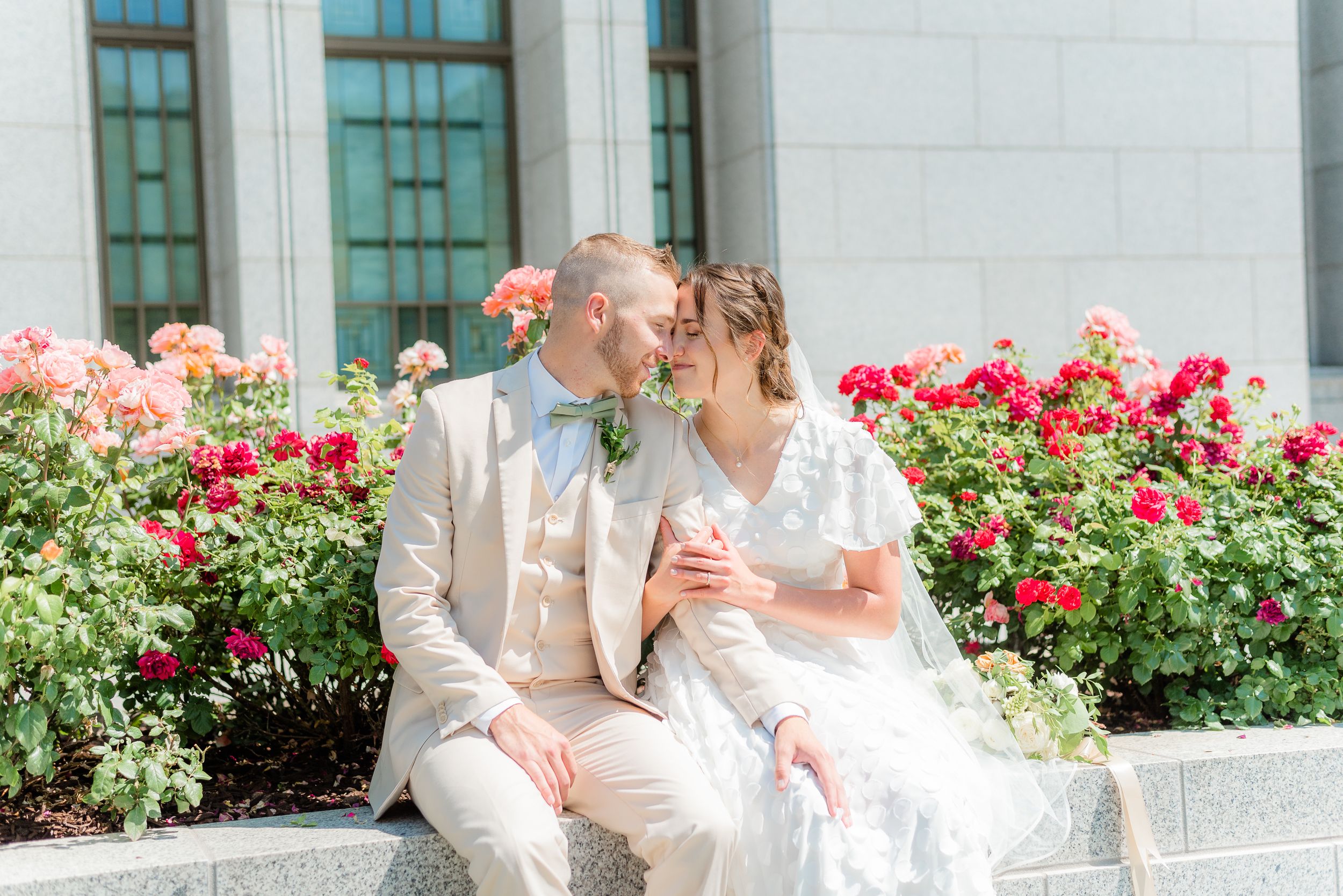 Bride and groom sit together sharing a sweet moment among vibrant red and pink roses Utah Draper Temple. Megan Snell Photo Utah Weddings