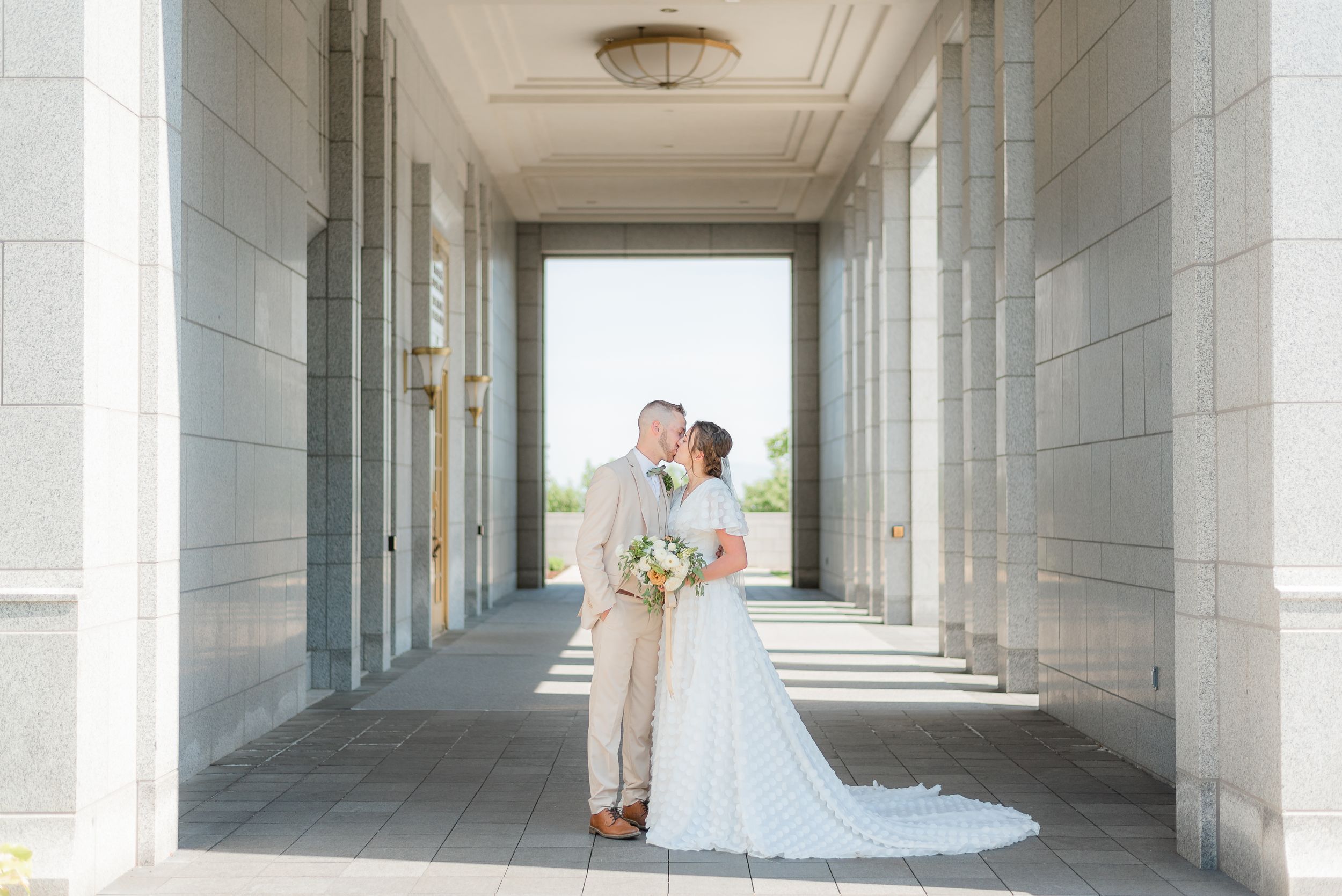 Bride and groom share a romantic kiss in a sunlit columned hallway on their wedding day. Megan Snell Photo Utah Weddings draper temple