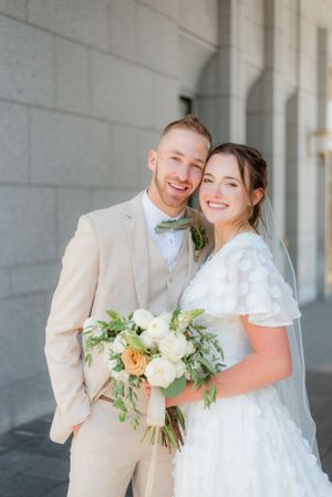 Smiling bride and groom pose together holding a white and green bouquet outside an LDS temple.