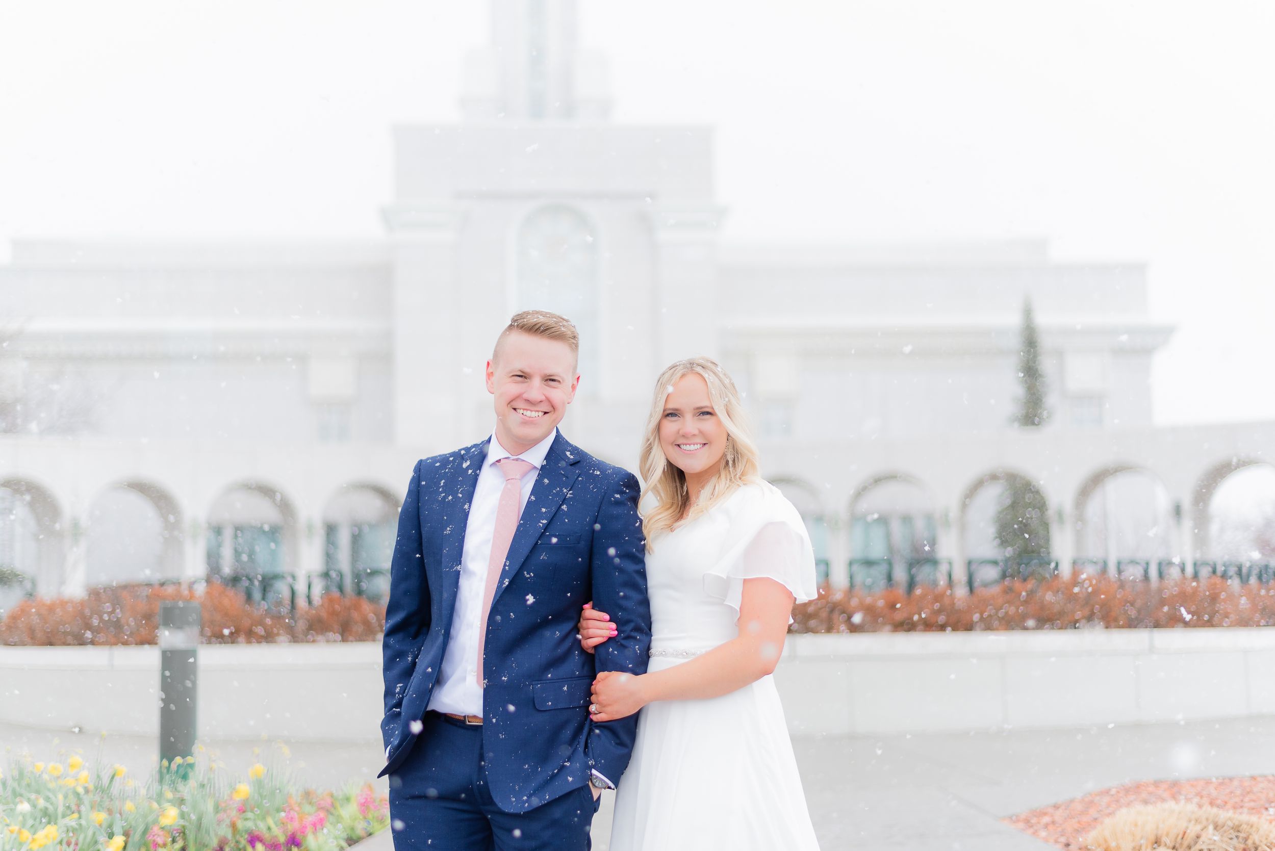Smiling bride and groom pose together outside a white temple in snowy winter weather. Megan Snell Photo Utah Weddings