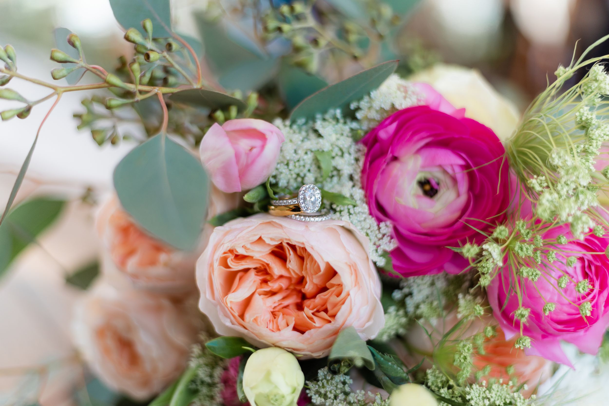 Close-up of a colorful wedding bouquet with pink ranunculus, peach roses, and a diamond engagement ring.