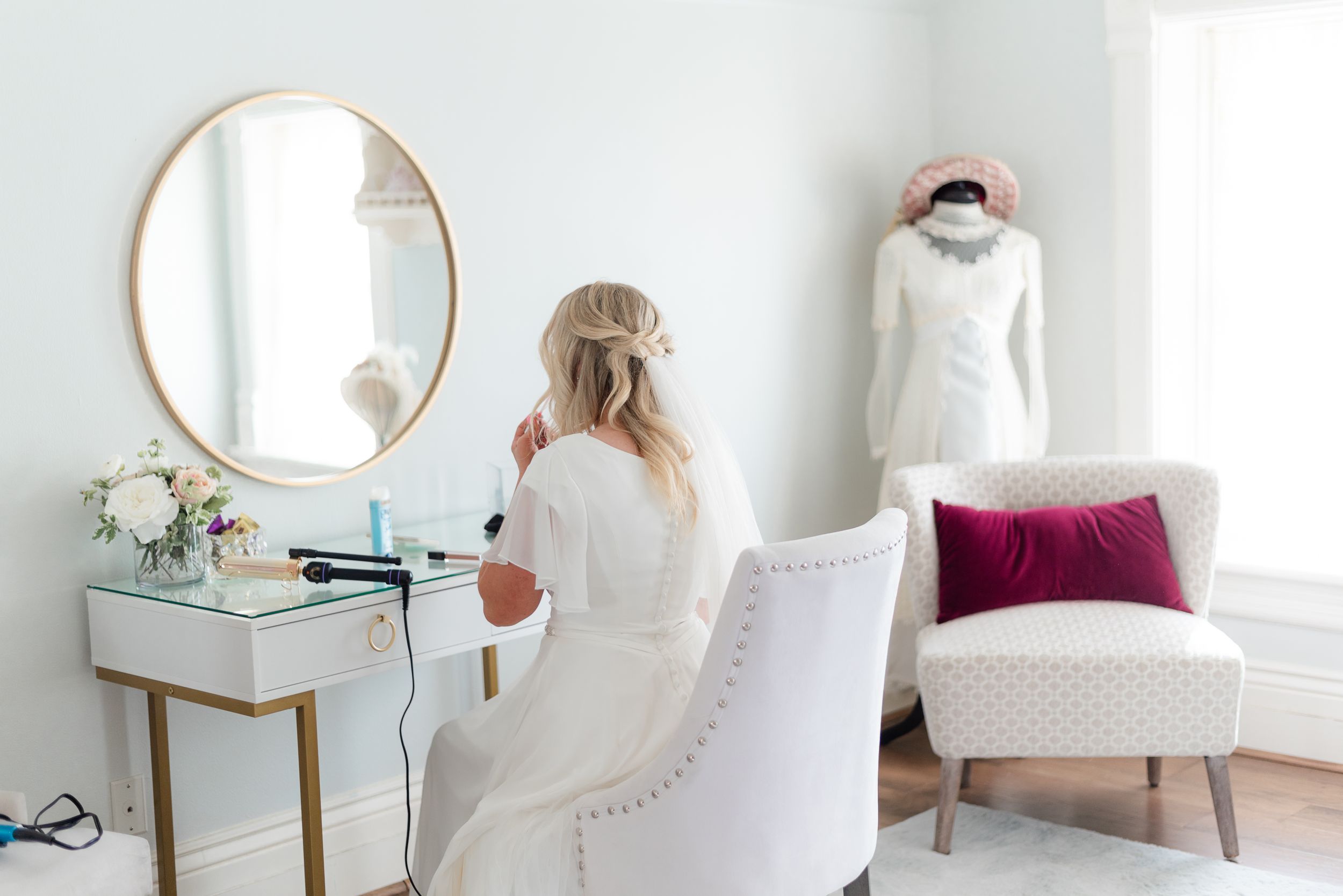 Bride sits at a vanity getting ready while her wedding dress hangs in a bright room. Megan Snell Photo Utah Weddings