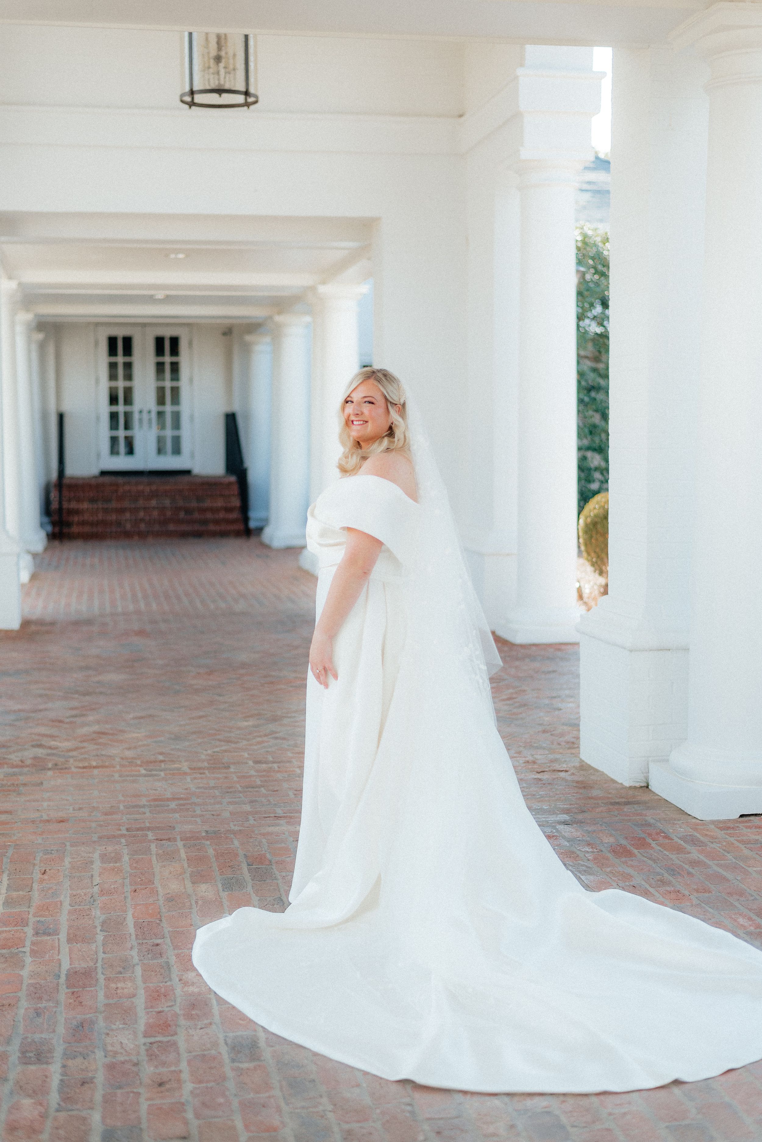 Elegant bride in off-shoulder white gown with long train posing between white columns on brick walkway. Megan Snell Photo Utah Weddings