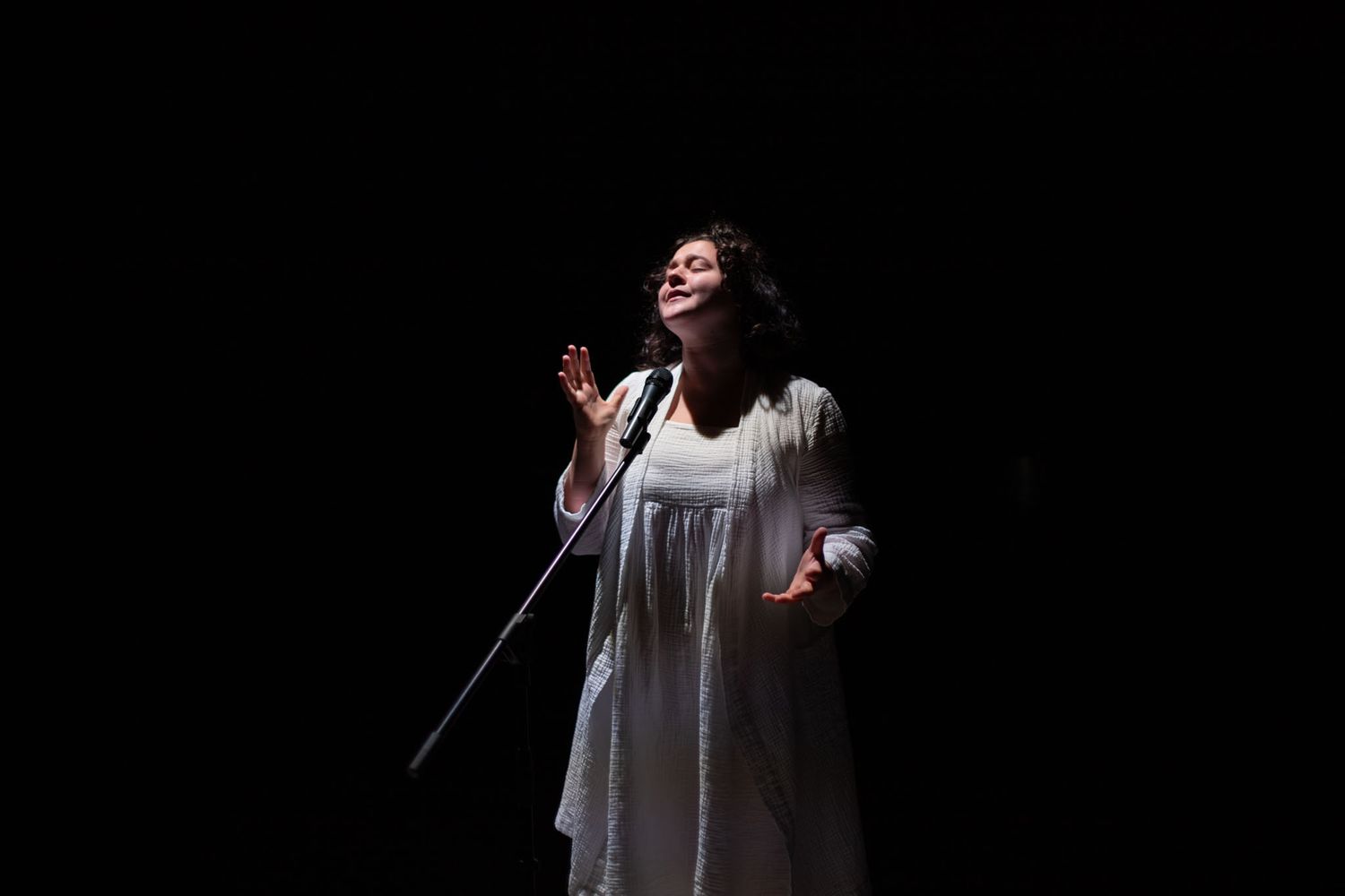 A performer in a flowing white dress sings on a darkened stage.
