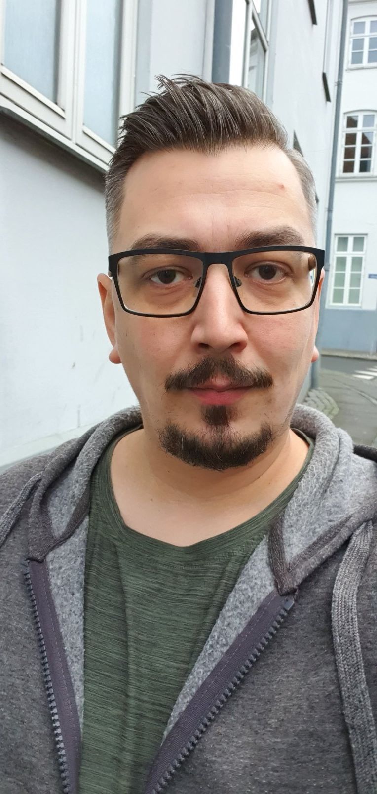 A young man with black rectangular glasses and a goatee wearing a gray hoodie takes a selfie outdoors.