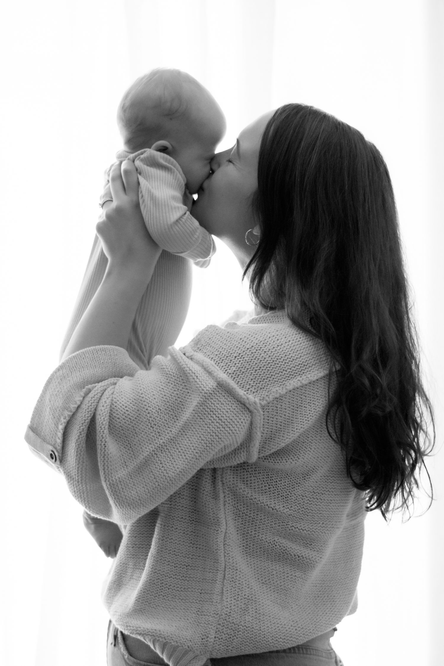 Black and white portrait showing tender moment between parent and infant in soft window light.