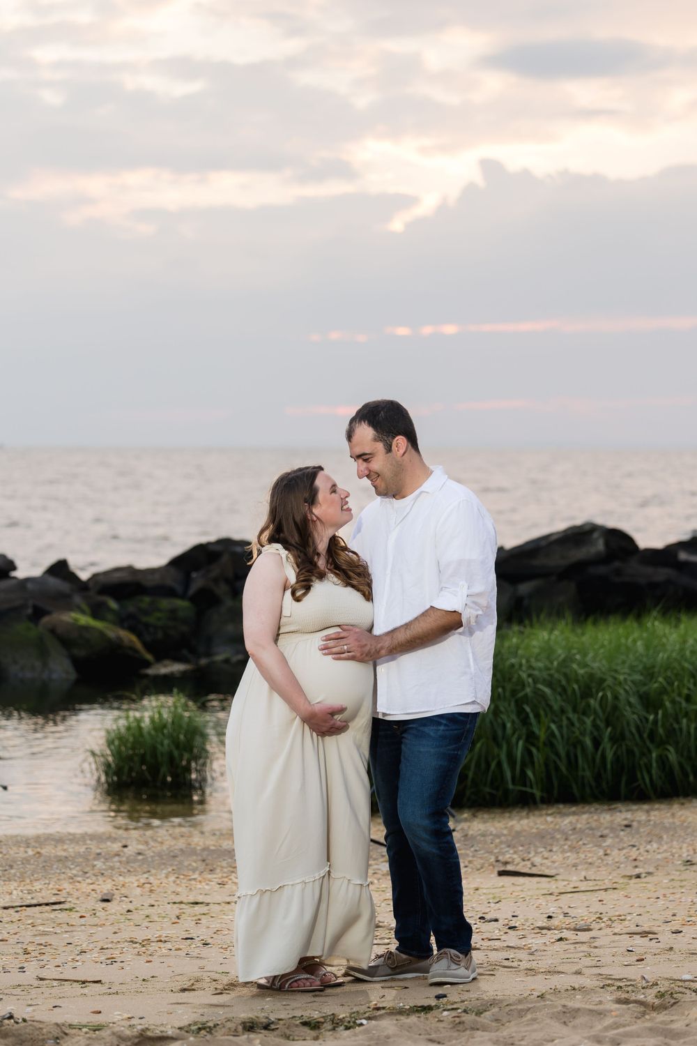 Couple poses for romantic sunset photos on beach with rocks and sea grass.