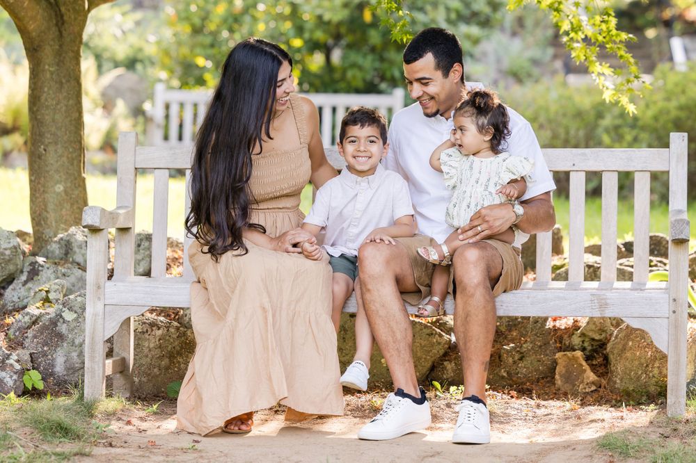 Family sitting together on white bench during outdoor photo session in golden sunlight.