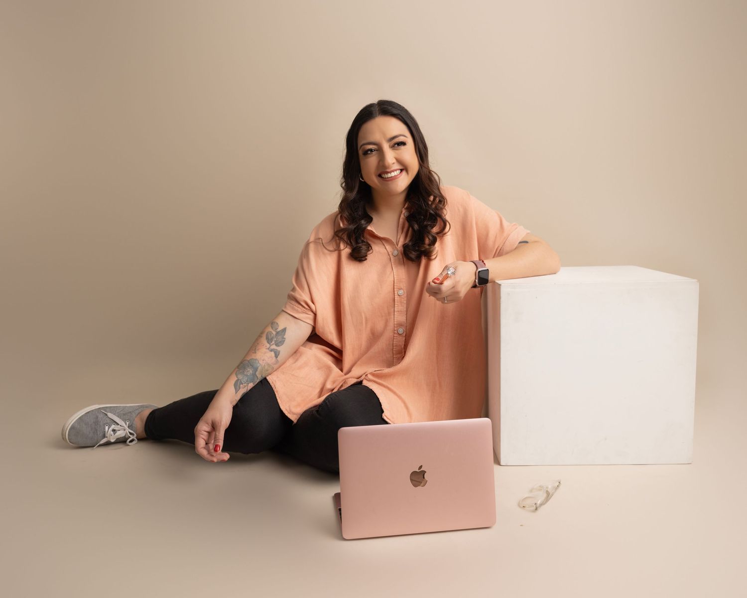 Lifestyle photo with person in peach sweater sitting by white cube with rose gold laptop.