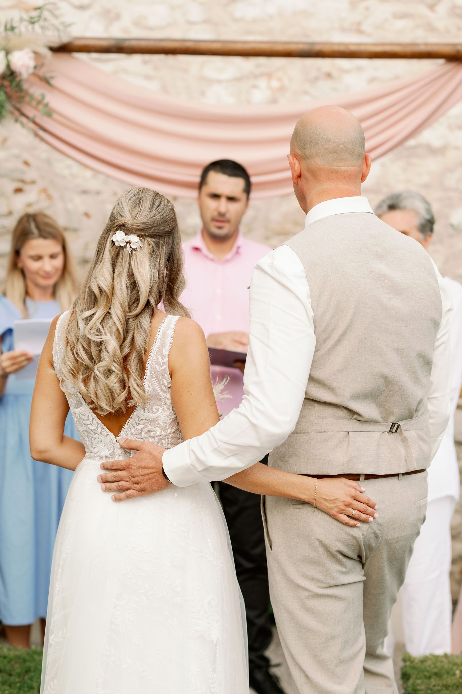 Wedding ceremony moment captured against stone wall with pink drapery and floral decor.