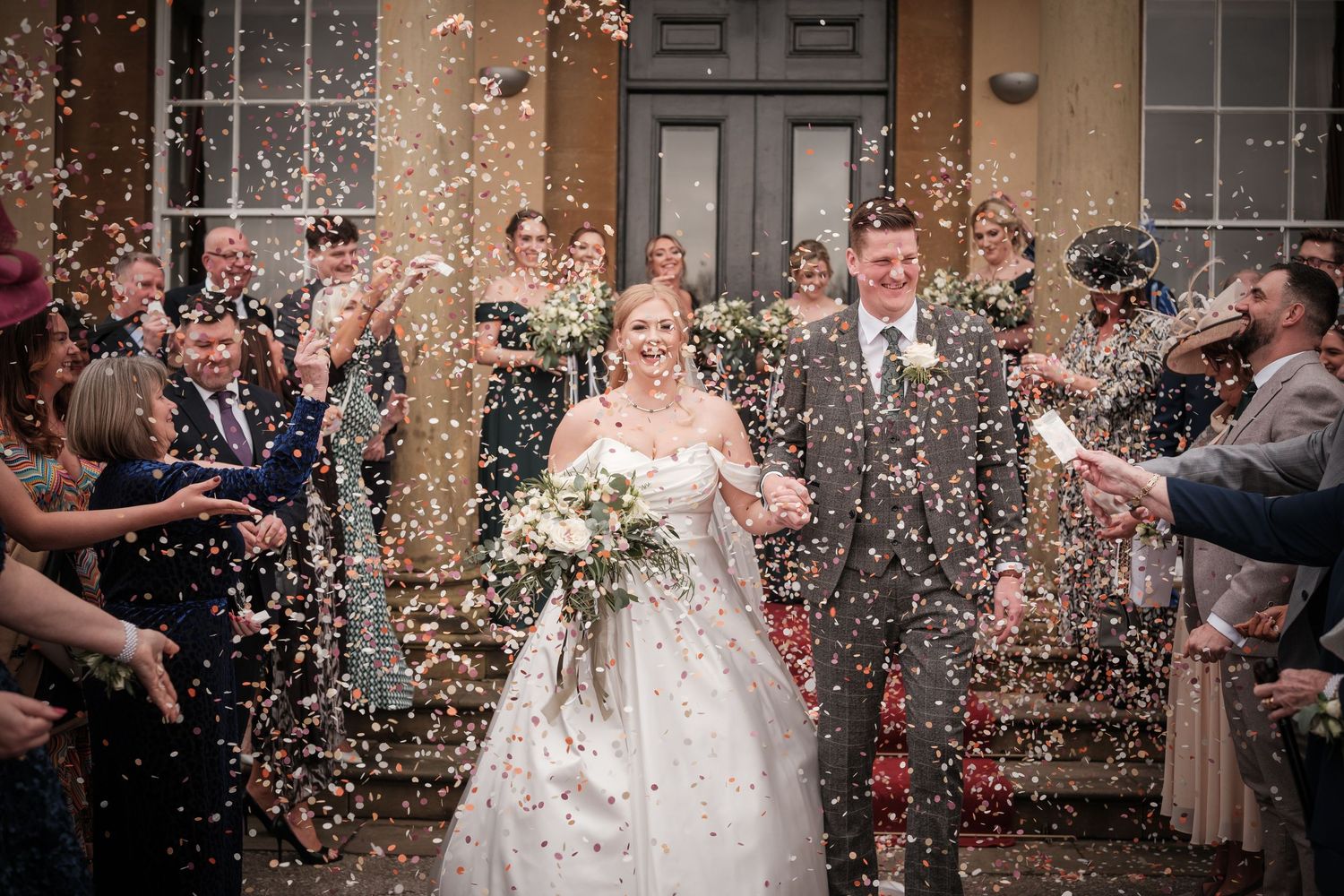 Wedding guests shower the newlyweds with confetti outside a historic building entrance.